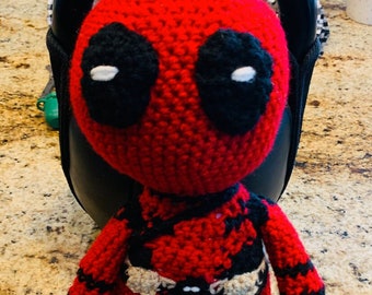 Op de afbeelding: Een gehaakte Deadpool-pop met een rood-zwart pak, witte ogen en een zwarte riem.