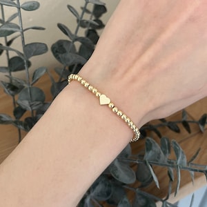 May include: A gold beaded bracelet with a small heart charm. The bracelet is on a person's wrist.