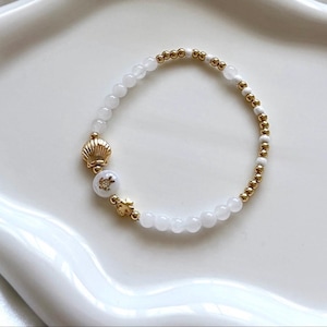 May include: A white and gold beaded bracelet with a seashell charm and a turtle charm. The bracelet is on a white surface.