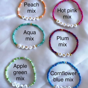 May include: Six beaded bracelets with different color combinations and names spelled out on each bracelet. The bracelets are peach mix with the name "ROBYN", hot pink mix with the name "BESTIE", aqua mix with the name "LEWIS", plum mix with the name "ENA", apple green mix with the name "NINA", and cornflower blue mix with the name "INESS".