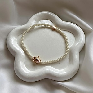 May include: A beaded bracelet with small, white beads and a gold-coloured turtle charm. The bracelet is presented on a white, flower-shaped dish, against a white fabric background.