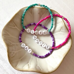 May include: Three beaded bracelets with the names Elsie, Marie, and Nat spelled out in white letters. The bracelets are layered on top of each other and are made with purple, pink, and turquoise beads.