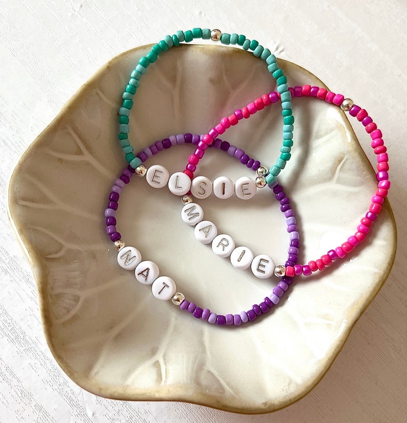 May include: Three beaded bracelets with the names Elsie, Marie, and Nat spelled out in white letters. The bracelets are layered on top of each other and are made with purple, pink, and turquoise beads.