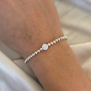 May include: A silver beaded bracelet with a white heart charm. The bracelet is on a person's wrist.