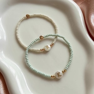 May include: Two beaded bracelets. One is white with gold accents and the other is light blue with gold accents. The light blue bracelet has a white charm with the letter "R" in gold. The second bracelet has a white charm with the letter "K" in gold.