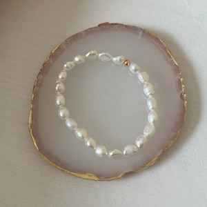 May include: A white pearl bracelet with a gold clasp. The bracelet is on a white and gold agate coaster.