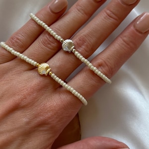 May include: Two beaded bracelets made of small, round, off-white beads. Each bracelet features a single metallic shell charm at the centre. One charm is silver-toned, and the other charm is gold-toned, with small metallic beads flanking each shell.