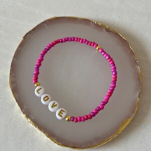 May include: A pink and purple beaded bracelet with gold accents and white beads spelling out the word "LOVE".