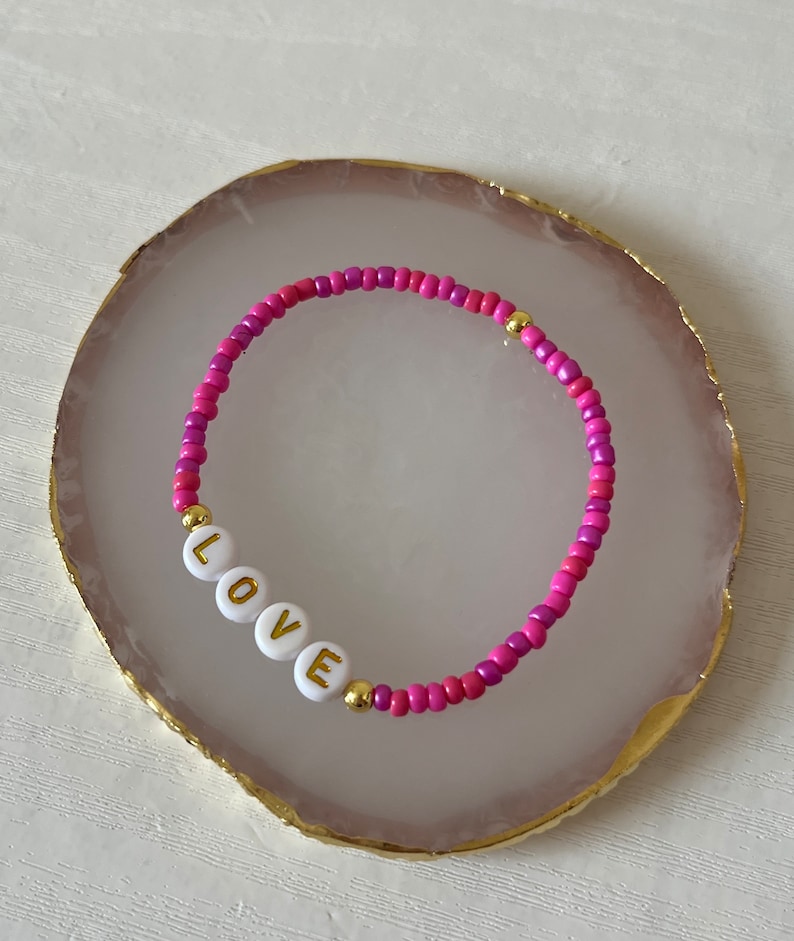 May include: A pink and purple beaded bracelet with gold accents and white beads spelling out the word "LOVE".