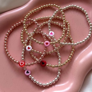 Può includere: Un set di cinque braccialetti con perline e ciondoli a forma di cuore. I braccialetti sono realizzati con perline dorate, oro rosa e argento. I ciondoli a forma di cuore sono rossi, rosa e bianchi.
