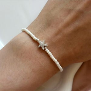 May include: White beaded bracelet with a silver starfish charm. The bracelet is made of small, round white beads and features a detailed starfish pendant in the centre. The bracelet is worn on a wrist.