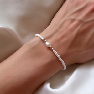May include: A white beaded bracelet with a single pearl accent. The bracelet is delicate and minimalist.