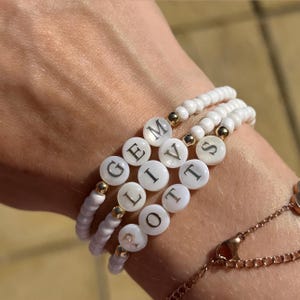 May include: Three white beaded bracelets with gold accents and lettered beads spelling out "GEM, LIV, and ROTT". The bracelets are stacked together, showcasing the personalized names. The bracelets are on a wrist.