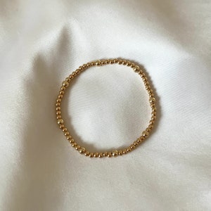 May include: Gold beaded bracelet. The bracelet is made of small, round, gold-coloured beads strung together to form a circular shape. The beads have a shiny finish and are evenly spaced around the elastic band.