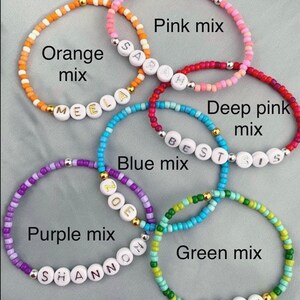 May include: Five colorful beaded bracelets with names spelled out in white beads. The bracelets are in shades of orange, pink, red, purple, and green.