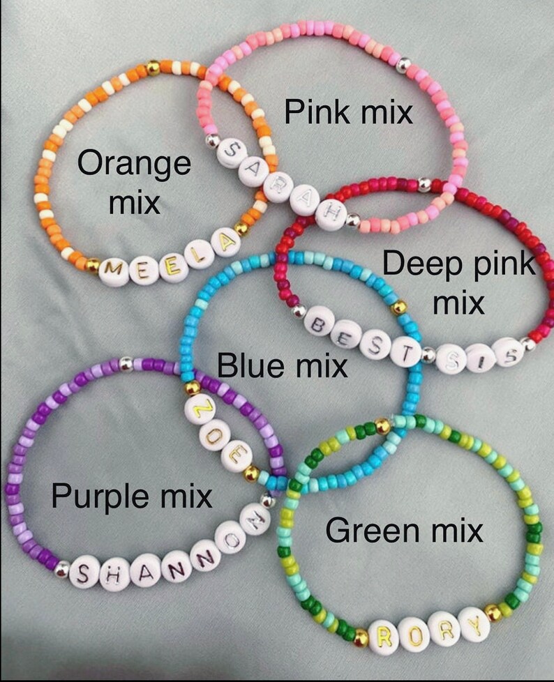 May include: Five colorful beaded bracelets with names spelled out in white beads. The bracelets are in shades of orange, pink, red, purple, and green.