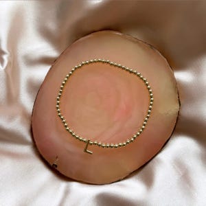 May include: Gold beaded bracelet with an "L" charm. The bracelet features small, round gold beads. The charm is a lowercase "L" in gold. The bracelet is presented on a pink and brown stone, with a silky, light pink background.