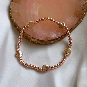 May include: A rose gold beaded bracelet with four larger rose gold discs. The bracelet is on a white surface.