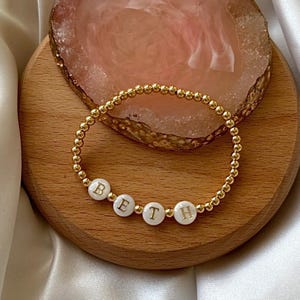 May include: A gold beaded bracelet with white beads that spell out the name "BETH".