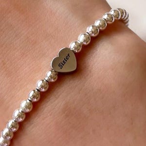 May include: A silver beaded bracelet featuring a heart-shaped charm. The charm is engraved with the word "Sister". The bracelet is composed of small, round, silver beads.
