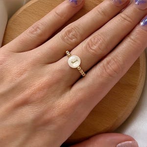 May include: A gold beaded ring with a white pearl-like centre featuring the letter "J" in gold.