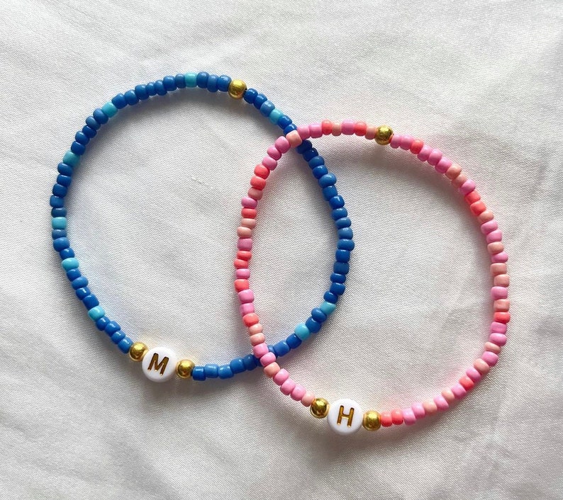 May include: Two beaded bracelets with gold beads and letter charms. The blue bracelet has the letter "M" and the pink bracelet has the letter "H".