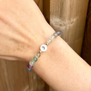 May include: A green, purple and white beaded bracelet with a gold charm featuring a crescent moon. The bracelet is on a person's wrist.