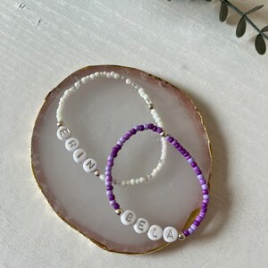 May include: Two beaded bracelets with the names "Erin" and "Bela" spelled out in white beads. The bracelets are on a pink and gold agate coaster.