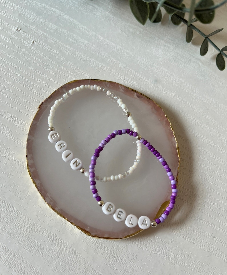 May include: Two beaded bracelets with the names "Erin" and "Bela" spelled out in white beads. The bracelets are on a pink and gold agate coaster.