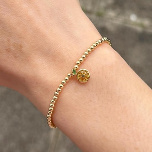May include: A gold beaded bracelet with a charm of a yellow lemon slice with green accents.
