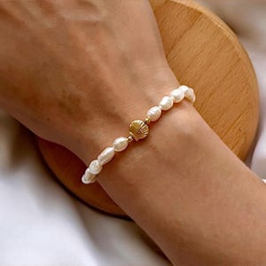 May include: A white pearl bracelet with a gold seashell charm. The bracelet is on a person's wrist.
