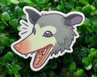 May include: A sticker of a cartoon opossum with a wide open mouth and large, white teeth. The opossum has gray fur and a white face with pink cheeks. The sticker is on a green background.