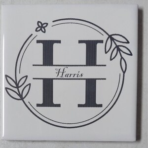 May include: White ceramic tile with a black and white monogram design. The letter "H" is surrounded by a circle with a floral design and the name "Harris" is written below the letter.