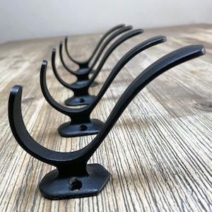 Puede incluir: Cinco ganchos de metal negro con un diseño curvo. Los ganchos están montados en una superficie de madera.