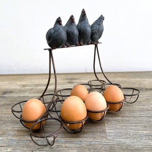 May include: A metal egg holder with six brown eggs and five black ceramic hens on top. The hens are arranged in a row, facing forward. The egg holder is on a wooden surface.