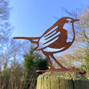 Könnte beinhalten: Eine rostige Metall-Robin-Silhouette, die auf einem Holzpfosten montiert ist. Die Robin ist nach rechts gerichtet und hat ein detailliertes Design mit einer strukturierten Oberfläche.
