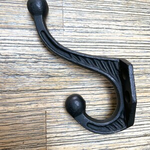 Puede incluir: Gancho de hierro fundido negro con un diseño decorativo de espiral y extremos redondeados. El gancho está montado en una superficie de madera.