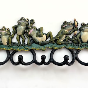 Puede incluir: Un colgador de pared decorativo de hierro fundido con cinco figuras de ranas verdes y crema sobre una base con forma de hoja. Las ranas están en varias poses, con ganchos negros debajo.