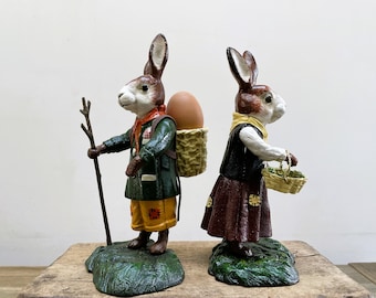 Puede incluir: Dos figuritas de cerámica de conejos de Pascua. Un conejo lleva una cesta con un huevo a la espalda. El otro conejo sostiene una cesta de flores. Ambos conejos están de pie sobre una base verde.
