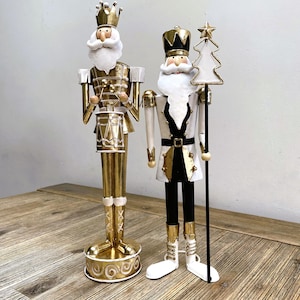 May include: Two decorative Christmas nutcrackers. One is gold-coloured, wearing a crown and holding drumsticks. The other is white, black, and gold, holding a staff with a star and Christmas tree design. Both have white beards.