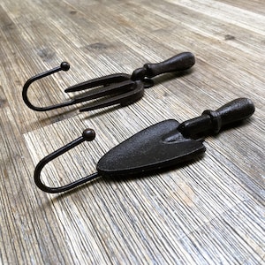 May include: Two black metal hooks shaped like gardening tools. One hook is shaped like a hand rake and the other is shaped like a trowel.
