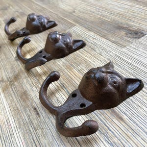 Può includere: Tre ganci a testa di gatto in ghisa marrone con ganci doppi. I ganci sono montati su una superficie in legno.