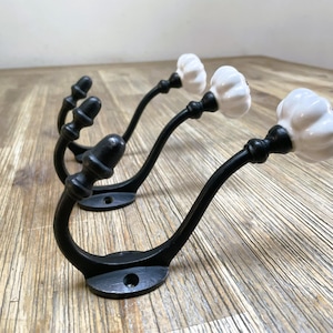 Könnte beinhalten: Set mit drei schwarzen Metall-Wandhaken mit dekorativen weißen Keramikknöpfen. Die Haken haben ein gebogenes Design und sind auf einer flachen Basis montiert. Geeignet zum Aufhängen von Mänteln oder anderen Gegenständen.