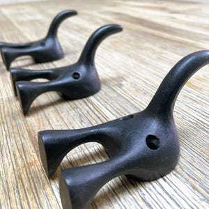 Może przedstawiać: Trzy czarne metalowe haczyki w kształcie stylizowanych zwierząt z zakrzywionym haczykiem na górze. Haczyki są zamontowane na drewnianej powierzchni.