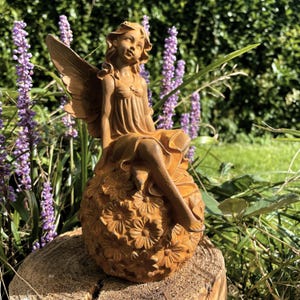 Può includere: Una statuetta di fata in metallo arrugginito marrone seduta su una sfera di fiori. La fata ha le ali e indossa un vestito. La statuetta è una decorazione da giardino.