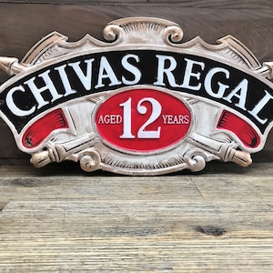 Könnte beinhalten: Ein silbernes und schwarzes Metallschild mit den Worten "CHIVAS REGAL" in weißen Buchstaben. Das Schild zeigt auch einen roten Kreis mit der Zahl "12" in weißen Buchstaben und den Worten "AGED" und "YEARS" in weißen Buchstaben.