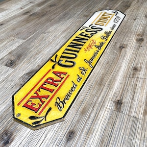 Könnte beinhalten: Ein rechteckiges Guinness Stout-Schild mit gelbem und schwarzem Design. Das Schild trägt die Aufschrift "EXTRA GUINNESS STOUT" in roter und schwarzer Schrift, darunter "Brewed at St. James's Gate, Dublin since 1759". Das Schild hat einen Vintage-Look.
