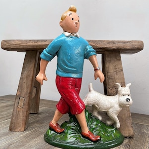 Könnte beinhalten: Figur einer Comicfigur in einem blauen Pullover und roter Hose, die mit einem weißen Hund geht. Die Figur steht auf einem grünen Sockel und vor einem Holzschemel.