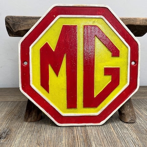 Op de afbeelding: Een rood achthoekig bord met een witte rand en een gele achtergrond. Het bord heeft de letters "MG" in rood.