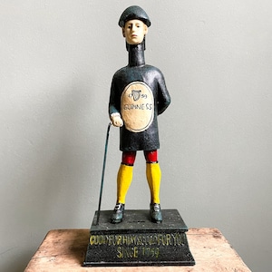 Pode incluir: Figura de publicidade Guinness vintage, pintada em cores escuras, com pernas amarelas, calções vermelhos e um casaco preto. A figura segura uma bengala e tem o logotipo da Guinness na frente. A base diz "GOOD FOR HIM & GOOD FOR YOU SINCE 1759."